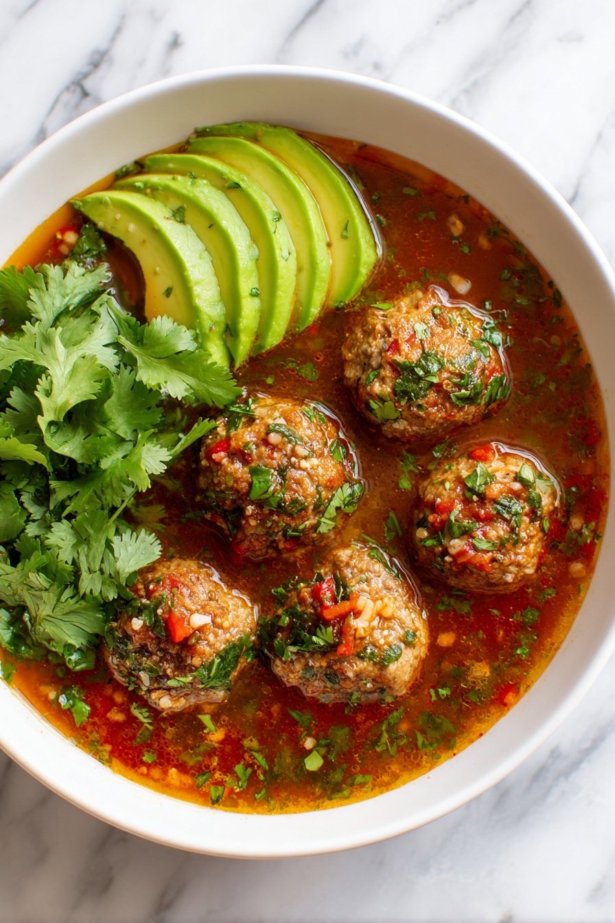 The image shows a white bowl filled with a rich, brownish-red broth that has visible pieces of diced red tomatoes and green celery floating in it. There are six round meatballs placed on top, each covered with finely chopped green herbs and small red pepper flakes. To the left side of the bowl, several slices of fresh avocado are neatly fanned out, showing their bright green color and smooth texture. A small bunch of cilantro leaves sits beside the avocado, adding more green to the dish. A silver spoon is resting inside the bowl on the right side. The bowl is placed on a white marbled surface. Photo taken with an iphone --ar 2:3 --v 7 - Albondigas Soup with Meatballs and Vegetables, Mexican Meatball Soup, Hearty Vegetable Soup, Easy Albondigas Recipe, Comforting Mexican Soup